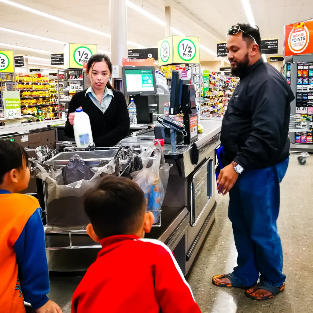 A father shopping with his two young children at an Australian supermarket checkout, capturing the everyday reality of family life and rising household costs.